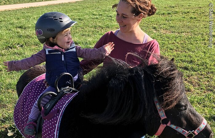 Ausritt auf dem Pony Ein Kleinkind sitzt auf einem schwarzen Pony. es wird von einer Frau mit braunen Haaren gehalten. Die Gruppe steht auf einer Wiese.