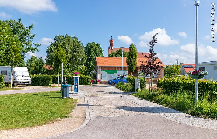 Wohnmobilstellplatz in Wolframs-Eschenbach Am Ende eines geplasterten Weges steht ein kleines Häuschen unter blauem Himmel. Gegenüber steht ein weißes Wohnmobil. Links und rechts des Weges sind grüne Sträucher und Wiese.