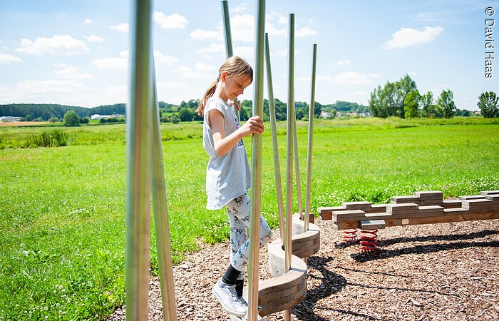 Motorikpark Ein junges Mädchen balanciert über einen Balken in Schlangenform. Dabei hält sie sich an Metallstangen fest. Im Hintergund grüne Wiesen.