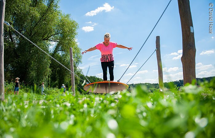 Motorikpark Windsbach Eine Frau balanciert auf einer runden Platte die von vier Seilen in der Luft gehalten wird. Die Seile sind an vier Holzpfosten befestigt die auf einer grünen Wiese stehen.