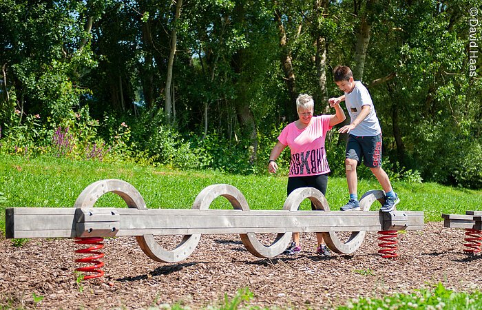 Motorikpark Windsbach Ein Junge balanciert über einen langen Holzbalken der auf zwei Sprungfedern befestigt ist. Eine Frau hält ihn an der Hand und stützt ihm beim Laufen über den Balken. Im Hintergund Wald und Wiese.