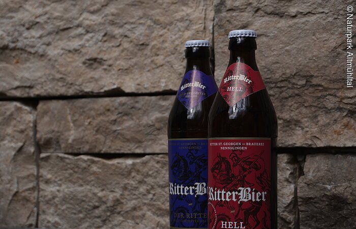 Bier Ritter St. Georgenbräu Zwei Bierflaschen mit Etiketten "RitterBier" vor einer Steinwand. Eine Flasche hat ein rotes, die andere ein blaues Etikett.
