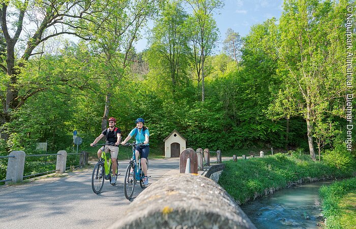 Anlautertal-Radweg bei Schafhausen Ein Pärchen ist mit ihren Fahrrädern auf dem Radweg unterwegs. Rechts begleitet sie die Anlauter. Der Wald strahlt in hellem grün. Hinter ihnen sieht man eine kleine Kapelle.