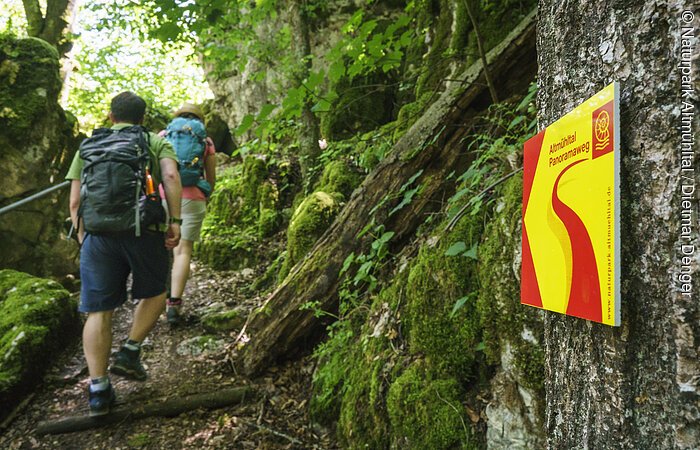 Im Vordergrund hängt ein Wanderschild des Altmühltal-Panoramaweges. Ein Wanderpärchen steigt die Treppen der Riedenburger Klamm hinauf.