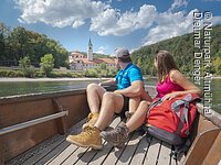Paar sitzt in einem Boot auf einem Fluss und blickt auf ein Gebäude mit Turm und Wald im Hintergrund.