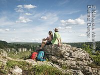 Zwei Wanderer sitzen auf einem Felsen und blicken auf bewaldete Hügel unter blauem Himmel mit Wolken.