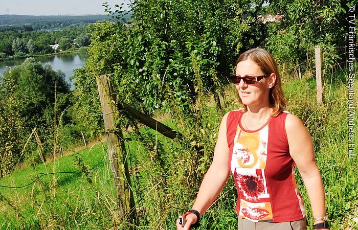 Eine Frau in sportlicher Kleidung und Sonnenbrille. Läuft mit Walking-Stöcken in der Hand auf einem Schotterweg entlang einer grünen Wiese. Im Hintergrund sind Bäume und Sträucher. In der Ferne sieht man einen See.