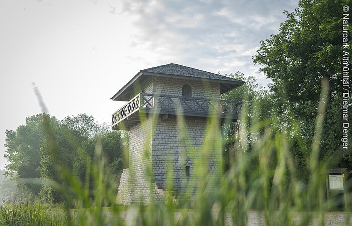Limesturm in Erkertshofen Der Limesturm ist aus weißem Stein rekonstruiert. Die Balustrade ist aus Holz und führt einmal um den ganzen Turm. Es ist unten ein kleines Fenster und oben eine Tür, die auf die Balustrade führt zu sehen. Umrahmt wird der Turm vom grünen Gras und den Bäumen im Hintergrund. Der blaue Himmel ist bewölkt.
