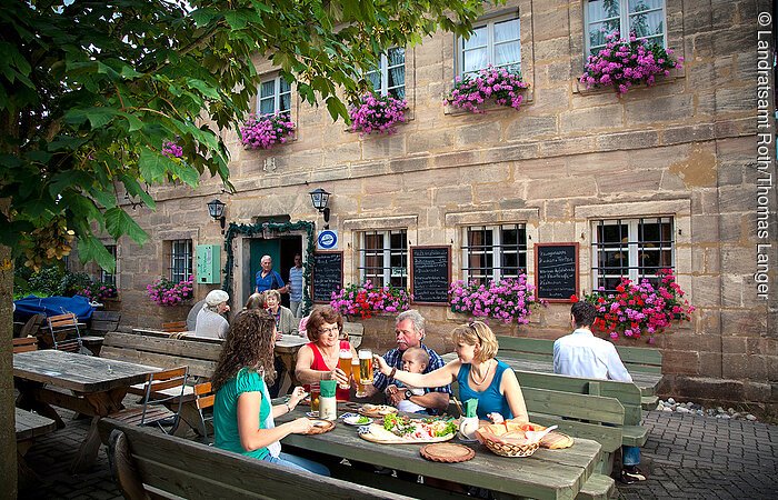 Bei der Brotzeit im Biergarten der Fuchsmühle bei Hilpoltstein stoßen Gäste mit kühlem Bier an und genießen die Köstlichkeiten. Die Gäste sitzen auf Sitzgelegenheiten aus Holz direkt vor dem Gasthof.