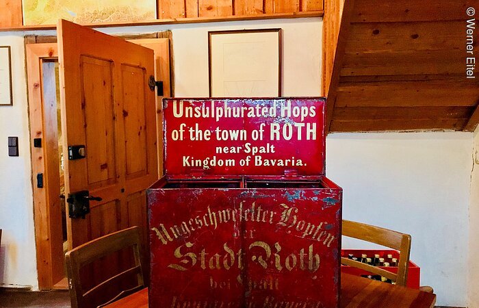 Eine rote Kiste aus Metall steht auf eibnem Tisch in einem Wohnraum. Auf der Kiste steht in weißer Schrift: Unsulphurated Hops of the town of Roth near Spalt Kingdom of Bavaria