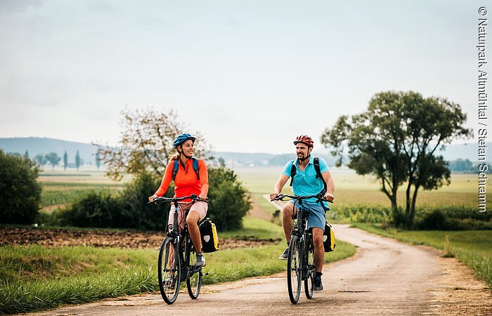 Eine Frau und ein Mann radeln auf dem Altmühltal-Radweg bei Dittenheim. Beide tragen einen Fahrradhelm und einen Rucksack. Der Radweg wird von Gras und grünen Bäumen gesäumt.