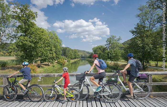 Vier Personen mit Fahrrädern auf einer Holzbrücke mit Blick auf einen Fluss und grüne Landschaft.