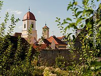Blick durch grüne Äste auf die Altstadt von Windsbach. Über den Häuserdächern ragt ein Kirchturm hervor. Ein Turm mit weiß-gelber Fassade und rotem Haubendach.