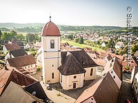 Luftbild einer Kirche umgeben von Häusern. Ein Kirchenschiff und Kirchturm mit heller oragener Fassade. Der Kirchturm hat ein Haubendach.