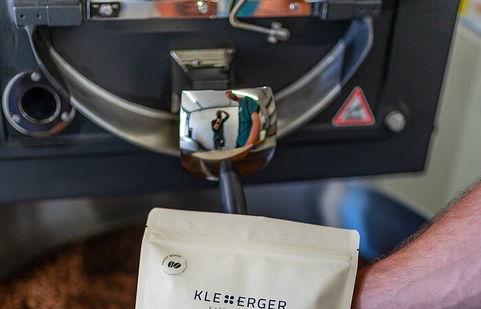 Kaffeerösterei Kleeberger Eine Hand hält eine Packung Kaffee in die Kamera. Dahinter eine große Maschine in deren Trommel Kaffeebohnen verarbeitete werden.