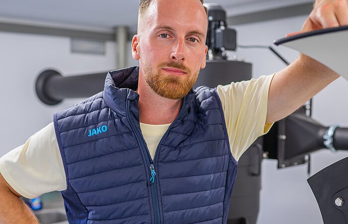 Kaffeerösterei Kleeberger Ein junger Mann mit blondem Haar und Bart lehnt an einer Maschine. Er trägt eine blaue Weste und blick entschlossen in die Kamera.