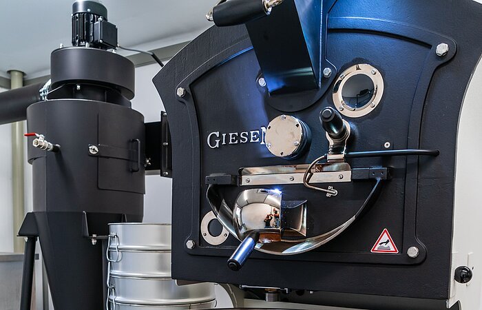 Kaffeerösterei Kleeberger Eine Kafferöstmaschine. Eine große weiße Blechtrommel mit einem Rüherer, der die Kaffeebohnen in der Trommel rührt.