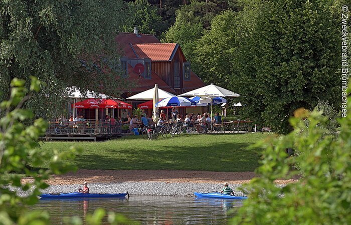 Kanufahrer auf dem See und der Kiosk Seeklause bei Absberg mit seinem barrierefreien Außenbereich Blick auf den See mit Kanufahrern und den Kiosk Seeklause Absberg mit Sitzmöglichkeiten und Sonnenschirmen am Ufer.