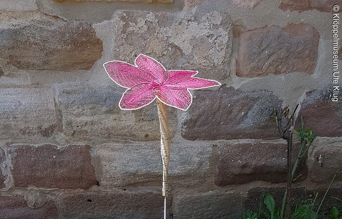 Eine geklöppelte rosa Blume steht in einer Grünfläche vor einer Mauer.