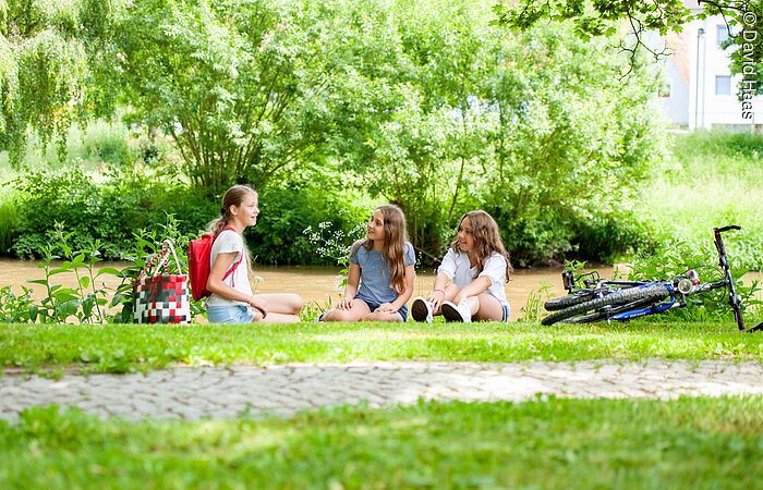 Drei junge Mädchen sitzen auf einer Wiese am Ufer eines Flusses. Sie unterhalten sich. Rechts von ihnen liegt ein Fahrrad. Im Hintergund fließt ein Fluss. Am gegenüber liegenden Ufer wachsen Bäume und Sträucher.