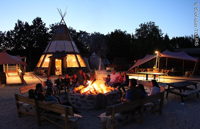 Lagerfeuerzeit Ein Gruppe Menschen sitzt in der Abenddämmerung auf Holzbänken um ein großes Lagerfeuer. Im Hintergrund stehen ein Tippi und ein Sonnensegel.