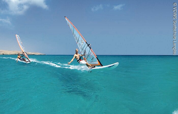 Zwei Windsurfer gleiten auf ihren Brettern. Das Seewasser ist türkis blau.