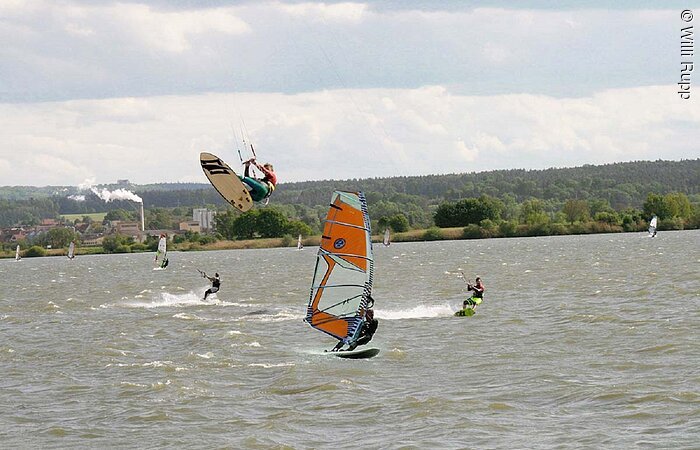 Surfschule Rupp Im Fokus ein Kitesurfer der über dem Wasser in der Luft hängt und ein Windsurfer.
Im Hintergund fahren weitere Kitesurfer über den See.