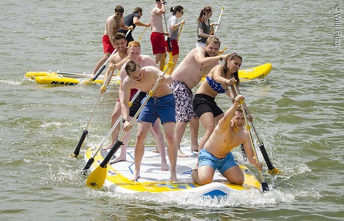 Paddeln Eine gruppe in Badekleidung steht auf einem großen Stand- Up Paddlingbrett und padelt durch das Wasser. Das Brett ist in etwas zwei mal drei Meter groß. Auf dem Brett stehen 6 junge Erwachsene. Dahinter eine andere Gruppe die über das Wasser paddelt.