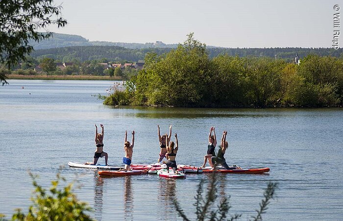 Yoga im Wasser Eine Gruppe von sehcs Sportlerin macht auf ihren Surfbrettern Yoga auf dem See. Die Bretter sind in einem Kreis angeordnet.