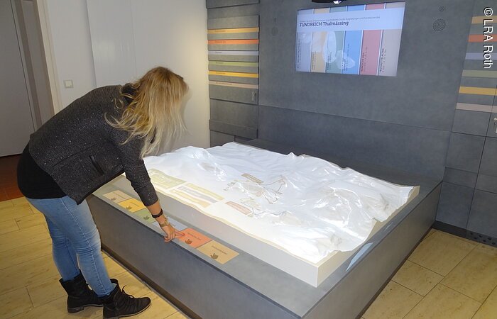 Eine Frau betrachtet ein Ausstellungstück in einem Museum. Ein 3D-Landschaftsrelief vor einer grauen Wand.