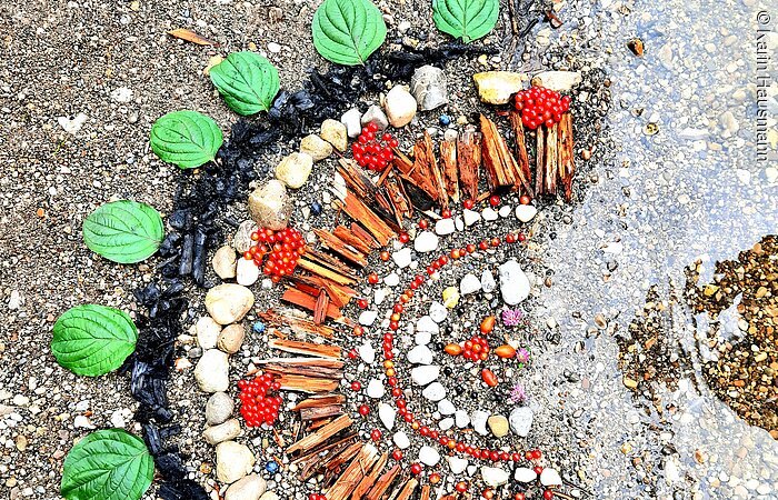 Nahaufnahme eines kreisrunden Mosaiks, gelegt aus Blättern und Steinen. Das Mosaik liegt direkt am Ufer eines Sees.