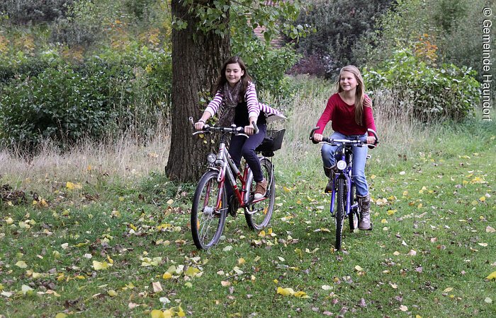 Zwei Mädchen radeln über eine grüne Wiese. Im Hintergund Bäume und Sträucher.