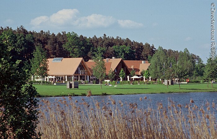 Direkt am See liegt der Gebäudekomplex des Starndhaus Birkach. Drei große Häuser mit Holzfassade an einer grünen Uferwiese. Dahinter ein großer Wald.