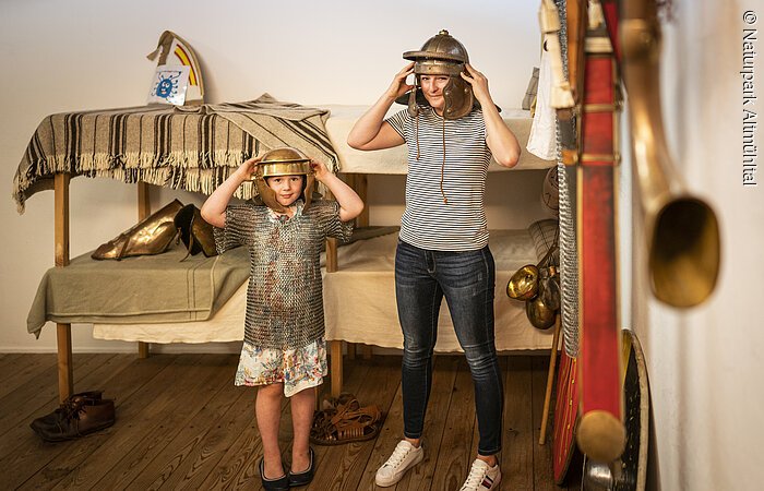 Kind und Frau stehen in einem Raum mit historischer Rüstung, beide tragen Helme und schauen in die Kamera.