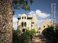 Steinernes Gebäude mit Rundturm und Weg mit Holzzaun bei sonnigem Himmel und Baum im Vordergrund