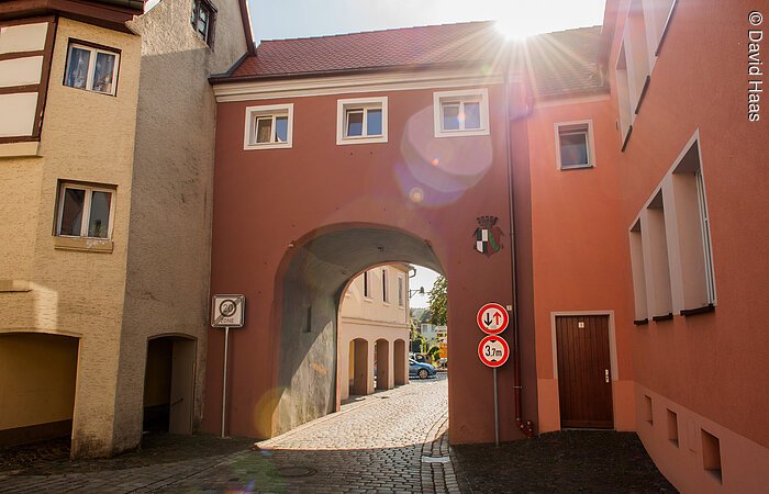 Ein Stadtor eingebunden zwischen zwei Häusern. Durch das Tor führt eine gepflasterte Straße der Altstadt. Über dem Torbogen drei kleine Fenster, dann folgt das Dach. Das Tor hat eine rötliche Fassade. Die Sonne spitzt über den Dächern hervor.
