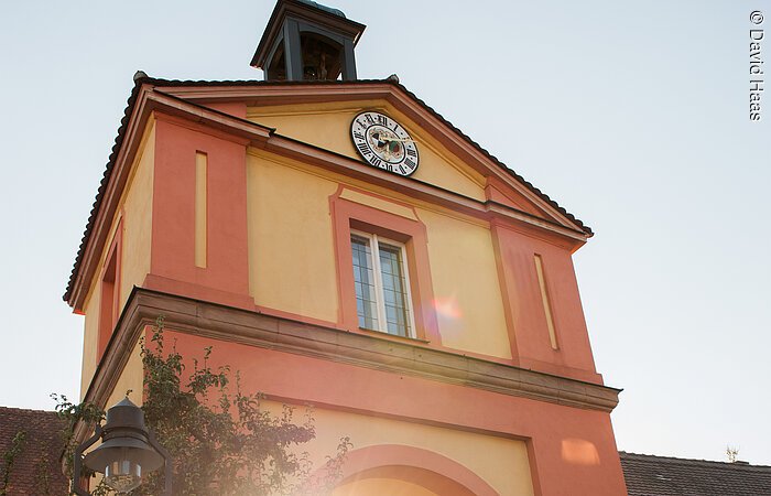 Ein Stadttor eingebunden zwischen einer Stadtmauer. Das Tor hat eine gelbe und rötliche Fassade. Ganz pben am Dachgiebel ist eine Uhr montiert. Durch das Tor führt eine gepflasterte Straße, außerdem scheint die untergehende Sonne durch das Tor.