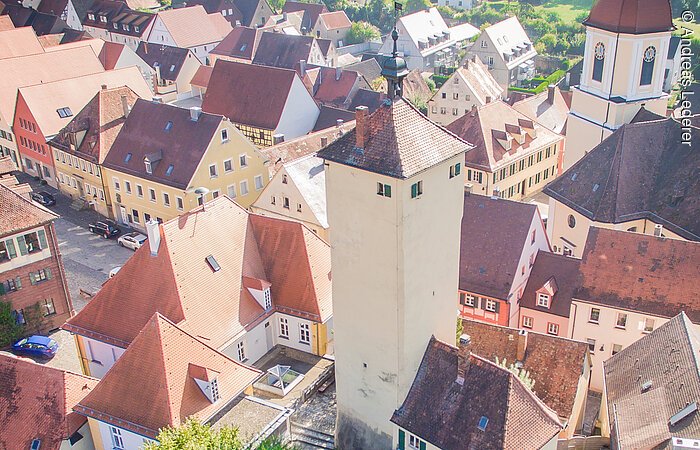 Luftbild eines Stadtturms. An den Turm angebaut ein kleines Häuschen mit grünen Fensterläden. Der Turm steht auf einer grünen Wiese umrandet von einer Mauer. Der Turm steht in einer Altstadt mit vielen Häusern.