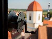 Im Fokus eine Spule mit Seil und Kurbel. Im Hintergund ist verschwommen ein Turm zu erkennen. Der Turm hat eine helle gelbe Fassade und ein rotes Haubendach.