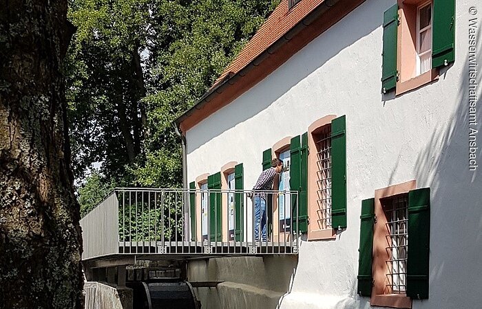 Mandlesmühle von außen. Ein großes weißes Haus mit grünen Fensterläden. Entlang des Hauses fließt ein kleiner Bach.
