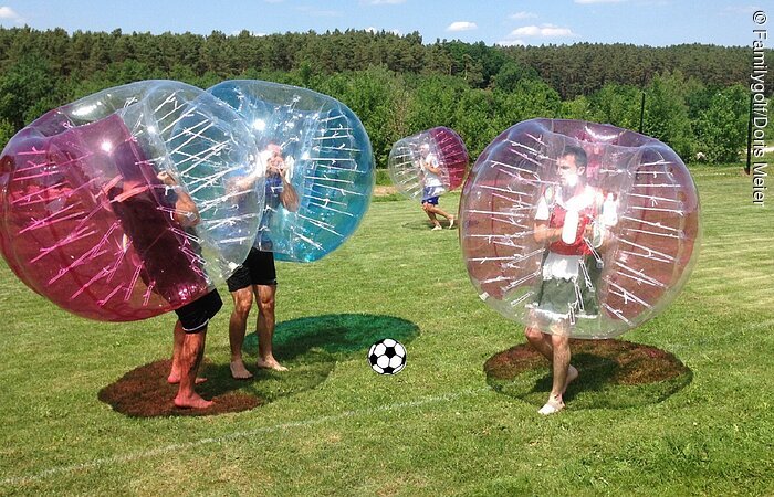 Bubble-Soccer in Pleinfeld Vier Menschen stehen spielen auf einem Fußballfeld und spielen Bubble-Fußball. Hierfür tragen die Spieler aufblasbare, transparente Kugeln über dem Oberkörper. Im Hutergrund stehen Bäume.