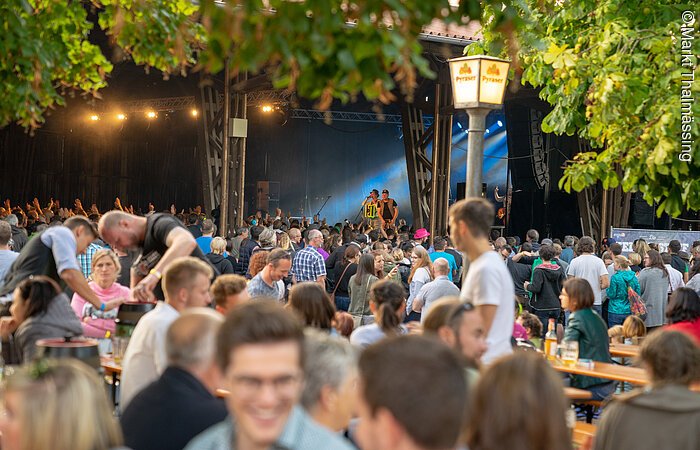 Ein voller biergarten. Viele Menschen sitzen auf Bänken an Tischen. Im Hintergund steht eine Bühne auf der gerade eine Band spielt.