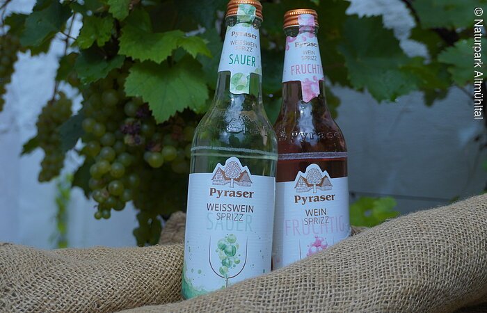 Zwei Flaschen Pyraser Weinsprizz, eine sauer, eine fruchtig, stehen auf einem Jutesack vor Weinreben.