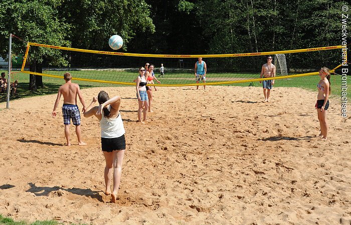 Jugendliche spielen auf einem sandigen Volleyballfeld. Dahinter beginnt ein Wäldchen.
