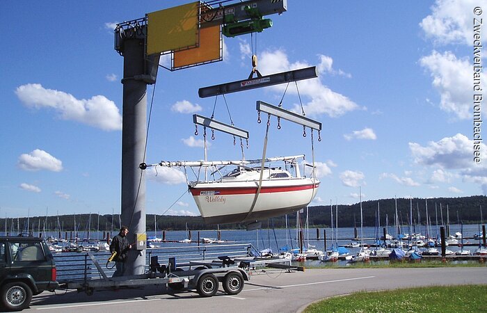 Einmal ganz anders Ein Segelboot wird mit der Hilfe Bootskran auf den Anhänger eines Autos geladen. Das rot-weiße Boot hängt in der Luft. Ein Mann bedient den Kran.
