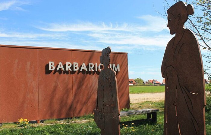 Barbaricum Burgsalach Zwei Frauenfiguren aus Metall ausgeschnitten stehen auf einer Wiese. Dahinter ein Metallbau mit dem Wort Barbaricum.