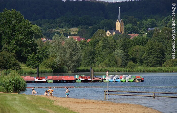 Pleinfelder Tretboote Blick auf den Brombachsee in Pleinfeld. Im Vordergrund sind badende Kinder am Strand, dahinter Trettboote am Liegeplatz zu erkennen. Im Hintergund ragt die Pleinfelder Kirche zwischen Bäumen hervor.