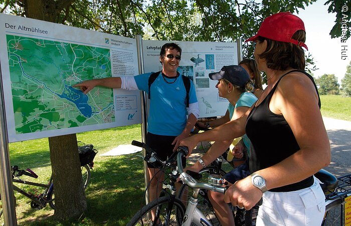 Radfahrer finden sich dank einer Info Tafel in Muhr am See und der Vogelinsel zurecht. Ein junger Mann zeigt mit ausgestrecktem Arm auf die Infotafel. Dabei lächelt er die drei jungen Frauen an, die auf die Karte blicken. Die Frauen halten ihre Fahrräder.