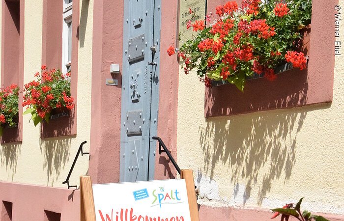 Eingang eines Hauses im fränkischen Baustil. gelbe FAssade mit roten Akzenten. Eine große graue Eingangstür aus Metall. Davor ein Schild mit dem Wort Willkommen und einem Foto eines Gartens mit grünem Gewächs.