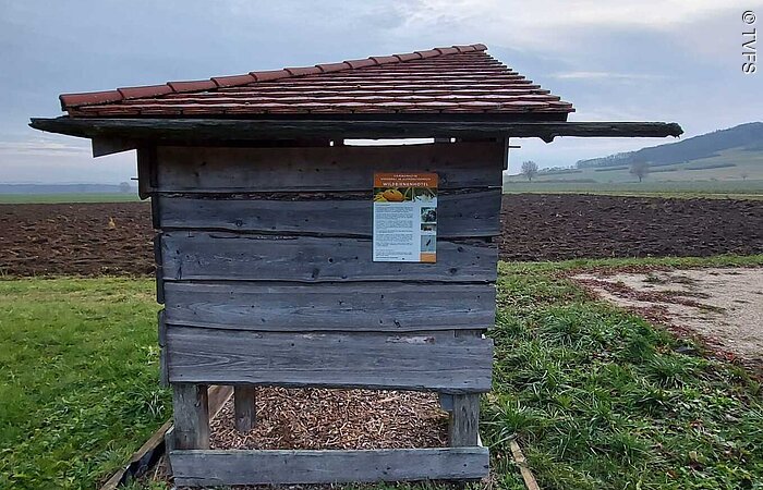 Kleine offene Holzhütte auf einer Wiese. Dahinter ein braunes Feld. Der Himmel ist bewölkt.